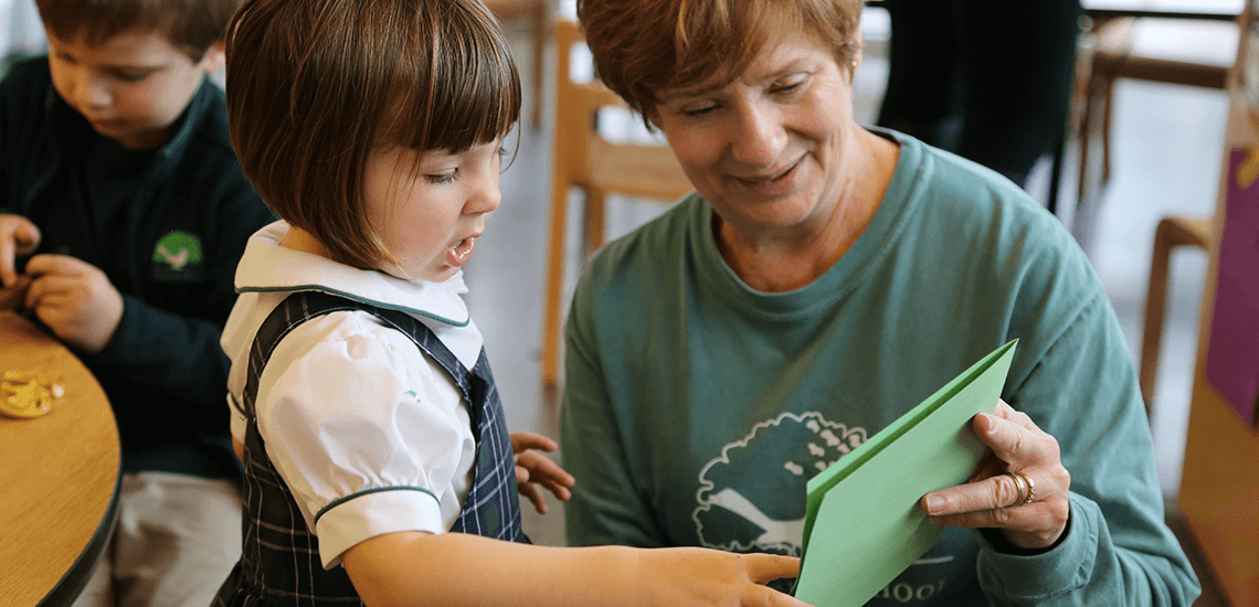 Teacher and young girl at The Parish School read a green paper together during a classroom activity, supporting early childhood communication and language development.